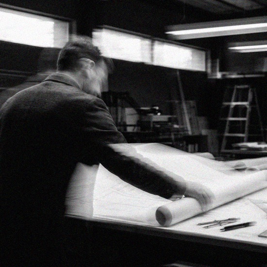 A person leaning over a drafting table, hands resting on rolled blueprints, in black and white.