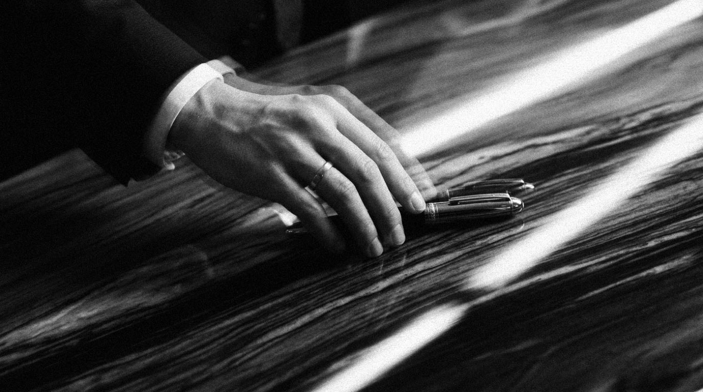 A hand picking up a fountain pen resting on a marble desk in warm light, in black and white.