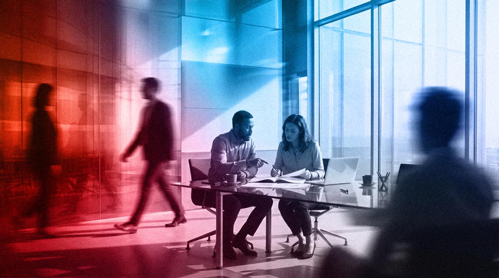 Two colleagues sitting together at a glass table in a high-rise office, reading a printed document between a laptop and an open notebook, while other people move past behind them.