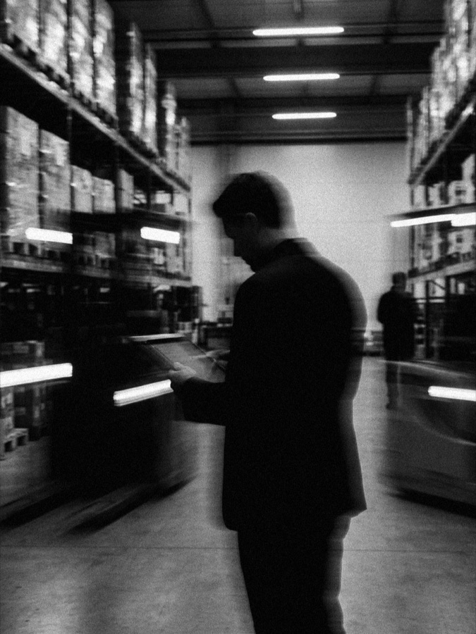 A warehouse worker in a pallet-racking aisle holding a handheld scanner, another figure with equipment beyond, in black and white.