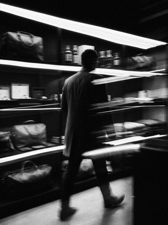 A person in motion through a boutique retail space lined with shelving of bags, bottles, and folded goods, in black and white.