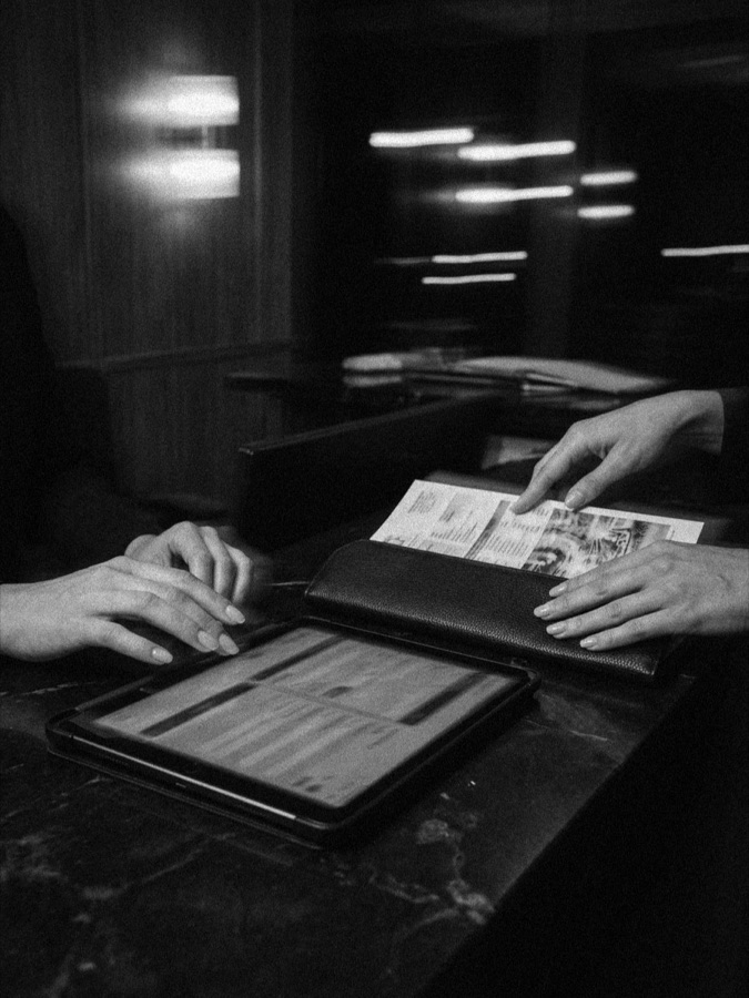 Two people's hands at a reception desk, one pointing to a printed document beside a tablet and a leather folio, in black and white.