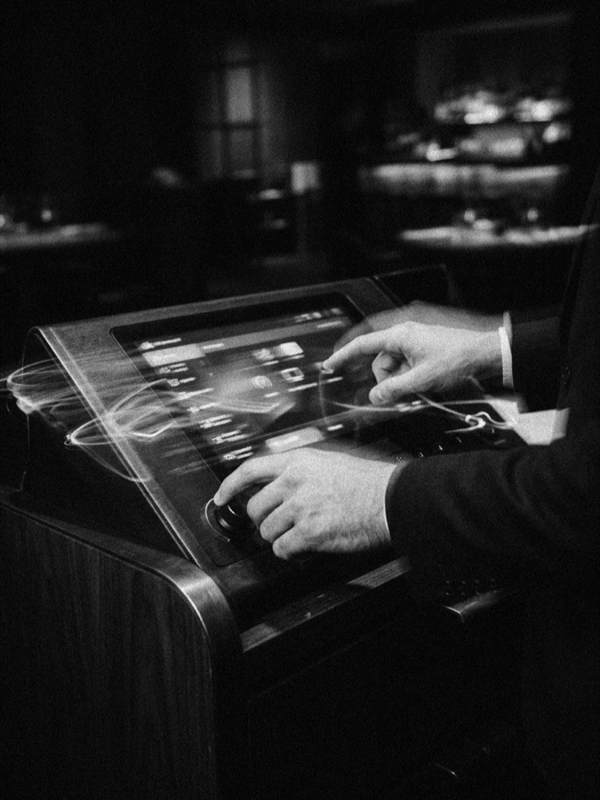 Hands adjusting a point-of-sale terminal in a moody bar setting, bottles lined on shelves behind, in black and white.