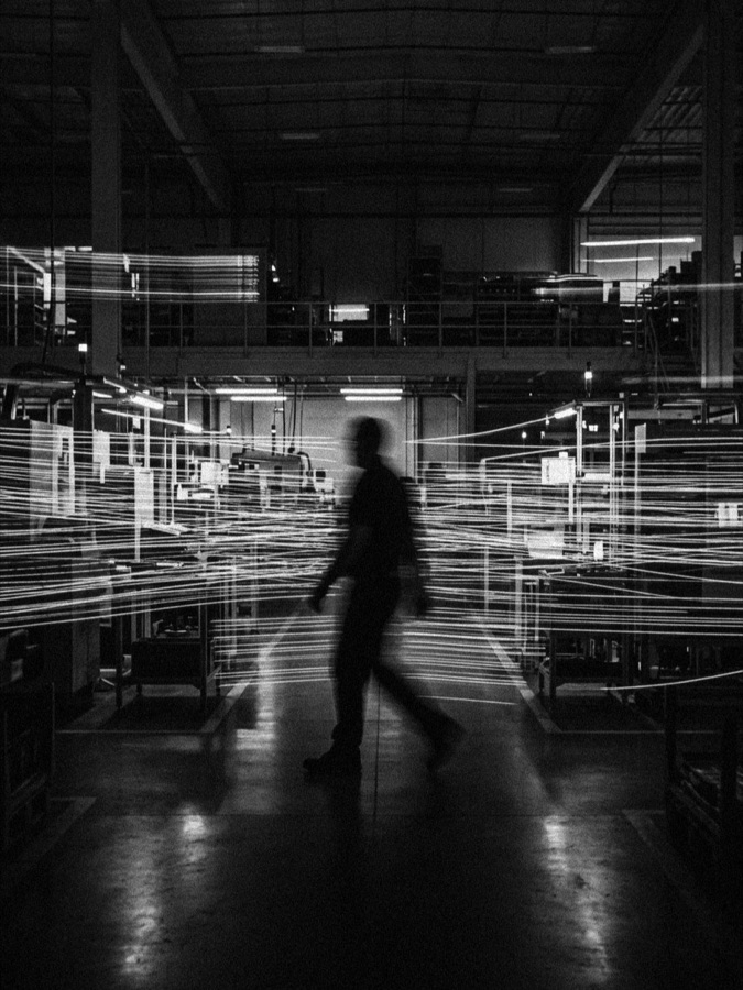 A supervisor walking a production floor past machinery and a mezzanine level, motion blur suggesting activity, in black and white.