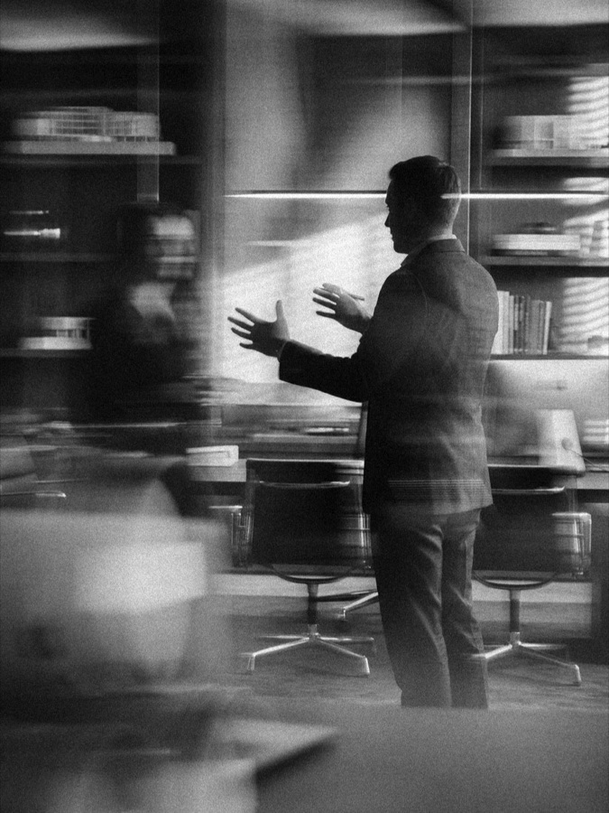A person in a suit gesturing mid-explanation in an office lined with bookshelves and architectural models, in black and white.