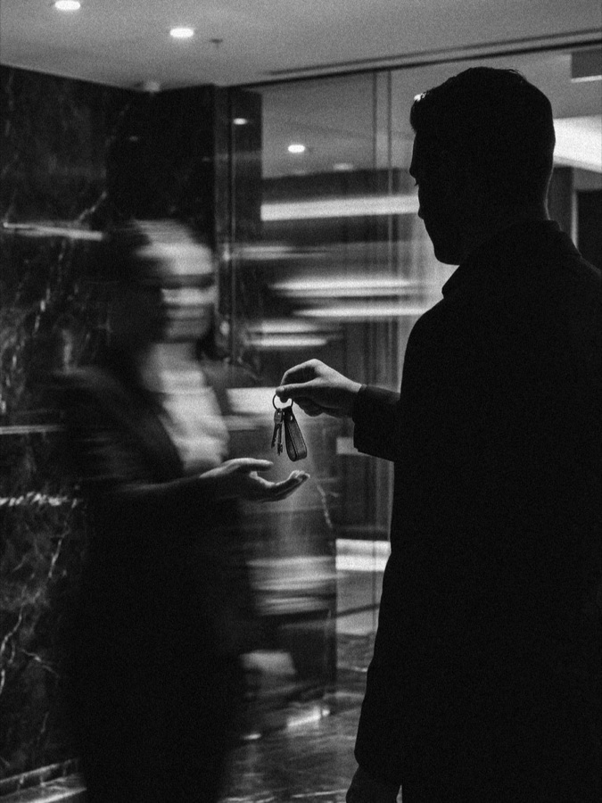 One person handing a set of keys to another inside a marble-walled lobby, in black and white.