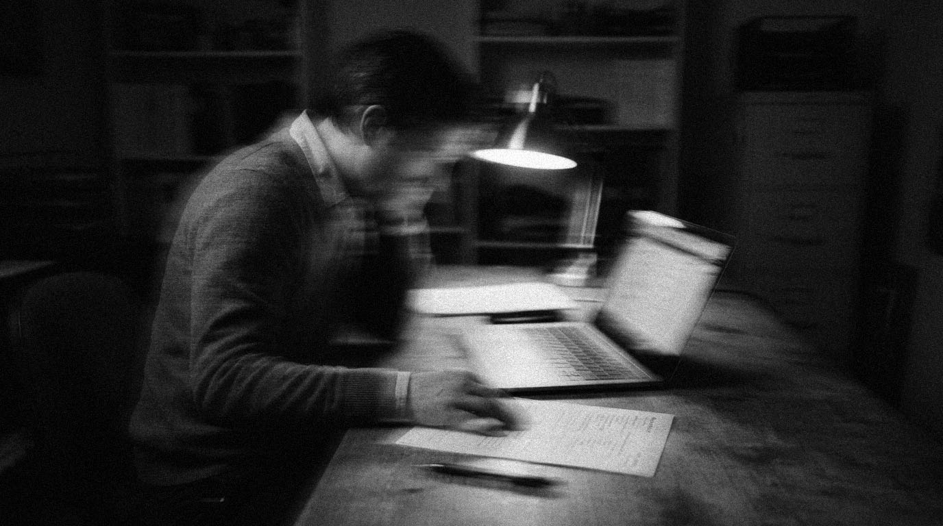 A business owner reading a printed worksheet beside an open laptop at a desk lamp late in the evening, in black and white.
