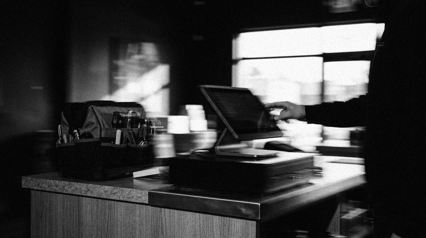 A hand adjusting a point-of-sale terminal on a small business counter beside an open tool bag, in black and white.