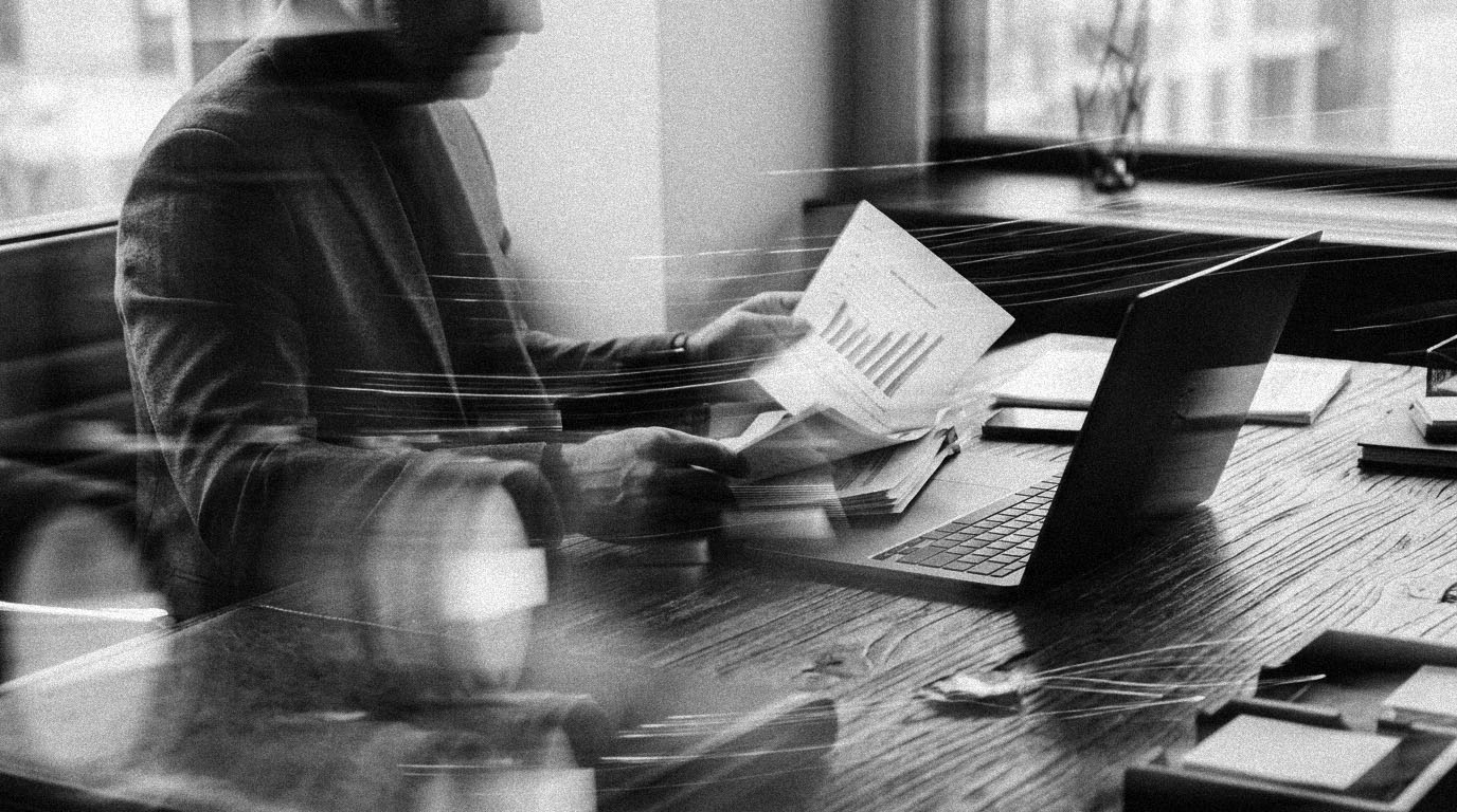 A manager at a desk holding a multi-page printed report open to a bar chart, a laptop beside it, in black and white.