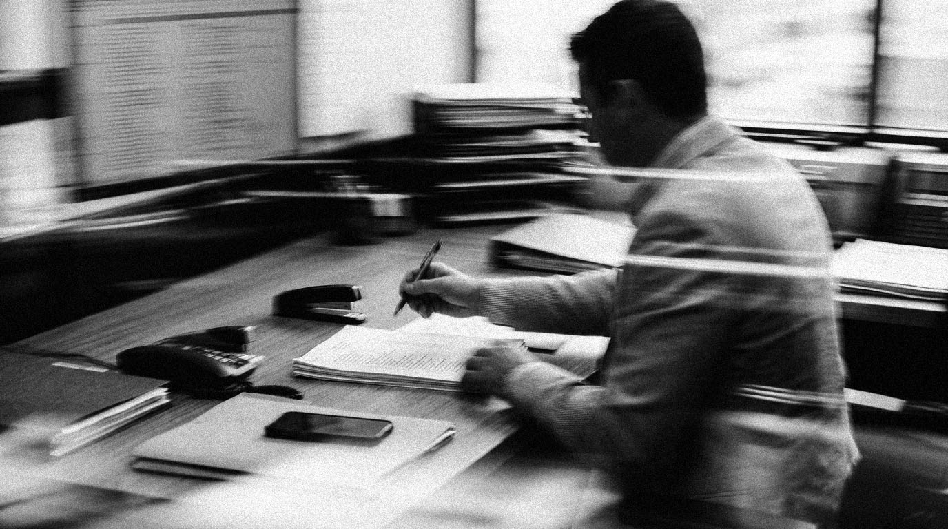 A staff member at a back-office desk checking items off a printed list with a pen, in black and white.