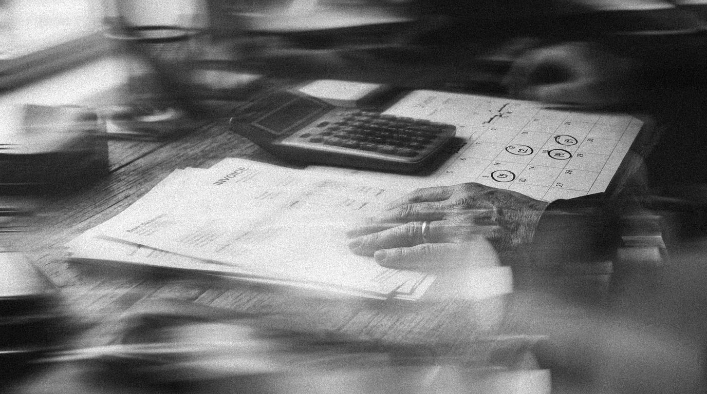 A hand resting on a stack of invoices beside a calculator and a calendar with several days circled, in black and white.