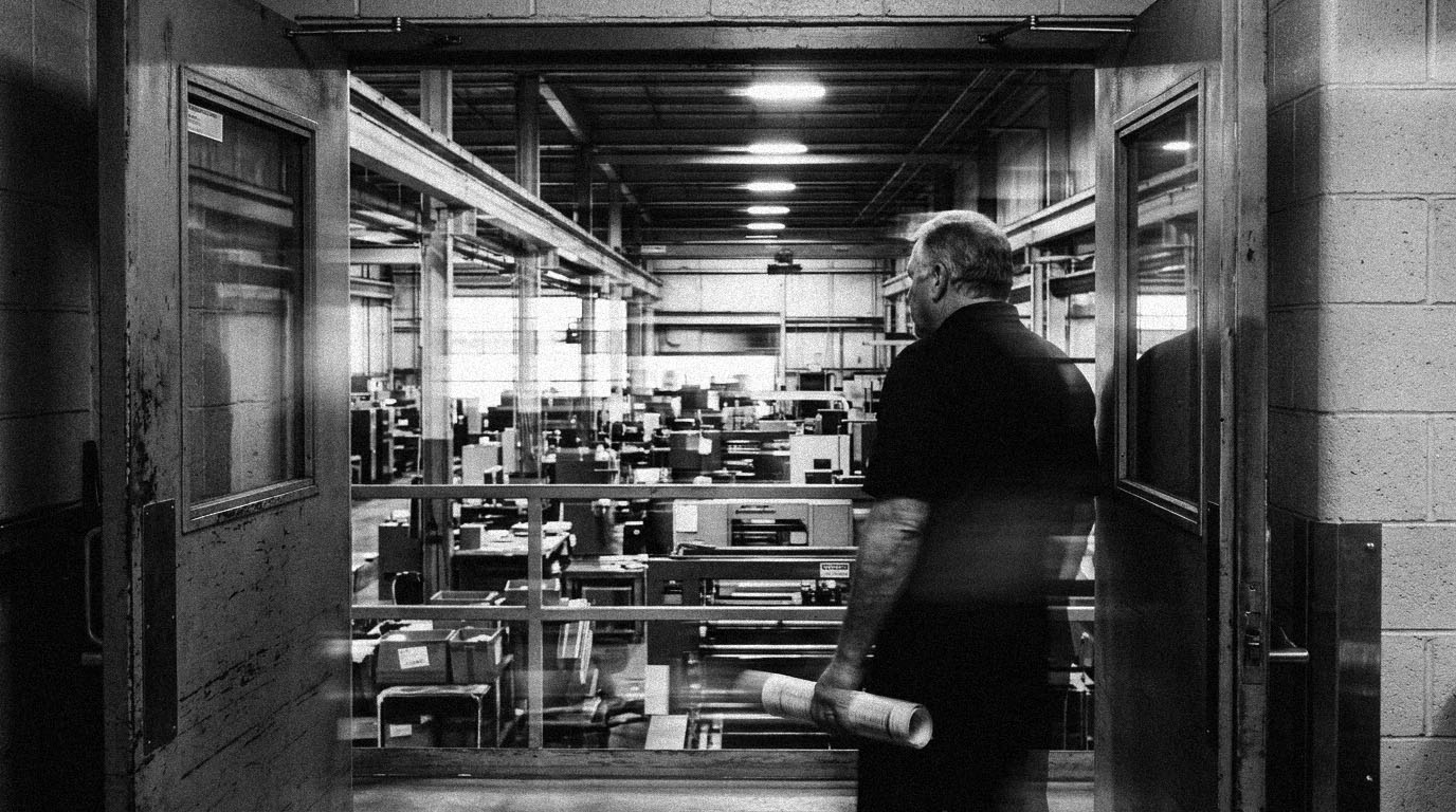 A senior operator in a manufacturing doorway looking out at the floor, a rolled report in hand, in black and white.