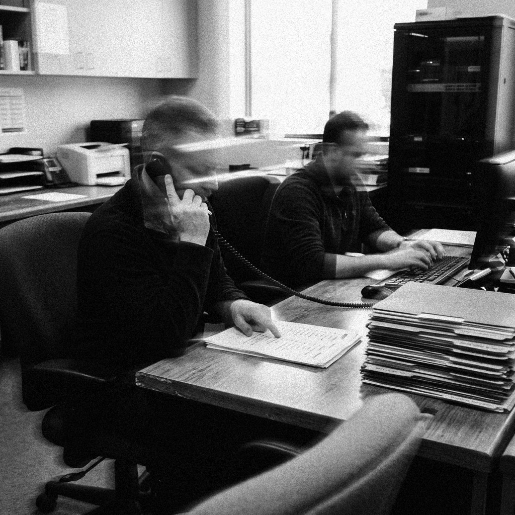 Two coworkers at adjoining desks in a back office, one on the phone and one at a keyboard, in black and white.